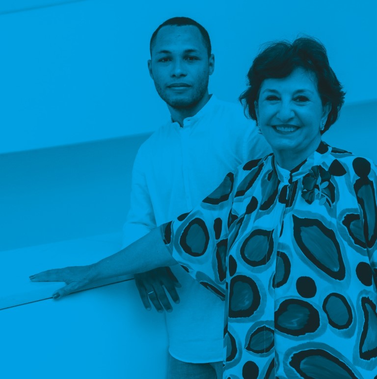 Descrição da imagem: fotografia horizontal colorida com filtro azul. Retrato de Santídio Pereira e Vilma Eidi. Ela está à direita da imagem com as mãos apoiadas em uma placa branca. Ela tem pele branca, cabelos curtos, lisos e escuro, usa blusa estampada de bolas concêntricas. Santídio está mais ao centro, atrás de Vilma, usa camisa branca de manga comprida. Ambos olham diretamente para a câmera. Fim da descrição da imagem.