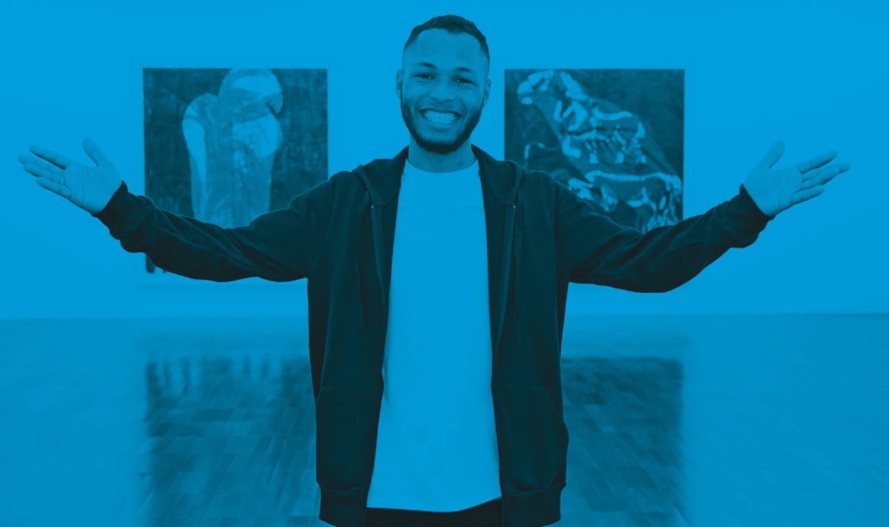 Descrição da imagem: fotografia horizontal colorida com filtro azul . Retrato de Santídio Pereira. Ele está de pé, de braços abertos e com um grande sorriso. Ele usa casaco preto, camiseta branca e está dentro de uma galeria com duas de suas gravuras de pássaros atrás de si. Fim da descrição da imagem.