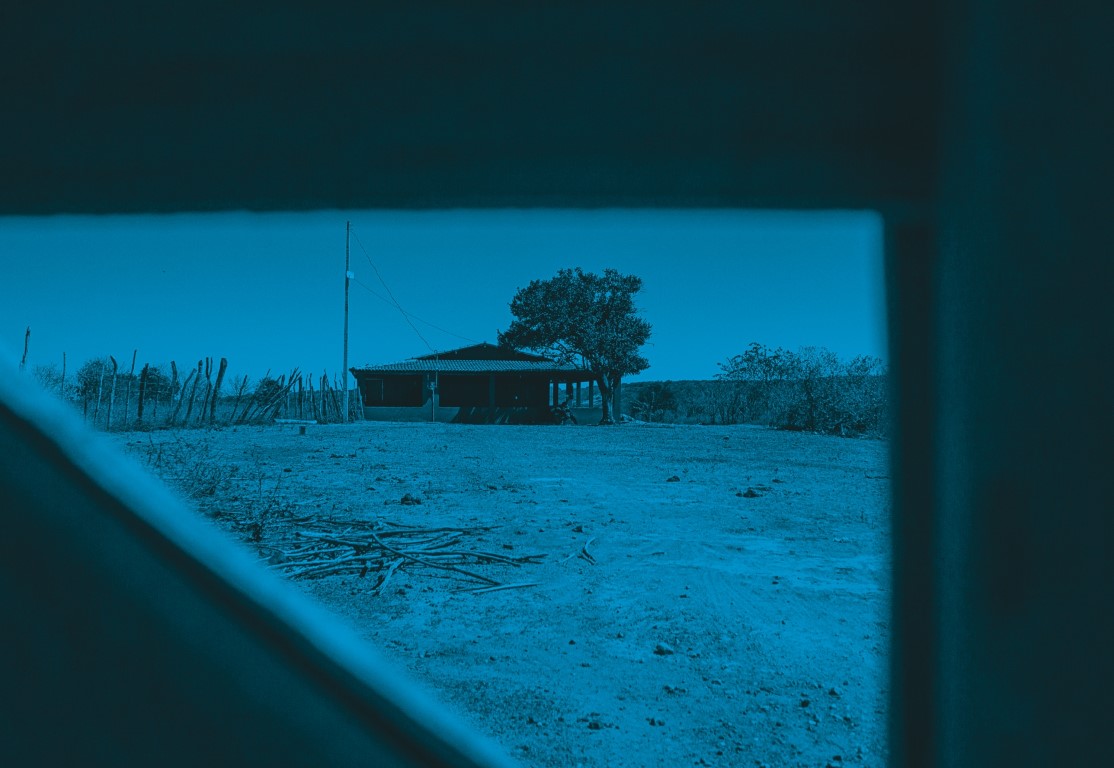 Descrição da imagem: fotografia horizontal colorida com filtro azul. Uma casa térrea de tijolos está centralizada na imagem. Tem telhado de telhas e varanda que se estende por toda a frente e lateral da casa. No lado esquerdo da casa há um poste de energia, e à direita há uma árvore que se debruça sobre o telhado da casa. À frente, uma área de terra batida com pilhas de galhos finos. A casa é vista através de uma fresta triangular de um portão, na qual emoldura a imagem. Fim da descrição da imagem.