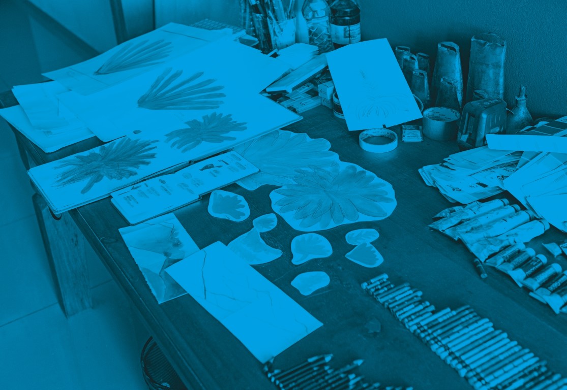 Descrição da imagem: fotografia colorida horizontal com filtro azul. Uma mesa de madeira com diversos papéis, cartões e objetos sobre ela. Há pelo menos quatro desenhos grandes de estudos de folhas e plantas sobre esses papeis. No centro há um caderno com desenhos coloridos de diversos objetos e seus nomes. Há diversos outros objetos sobre a mesa, como recortes de papeis no formato das plantas dos estudos, fita crepe, grafite, potes de tintas, lápis de cor, giz pastéis, entre outros. Fim da descrição da imagem.