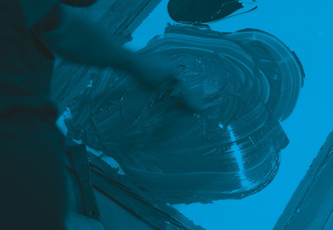 Descrição da imagem: fotografia horizontal colorida com filtro azul. Uma pessoa está com a mão direita espalhando uma tinta sobre uma placa, de forma circular. Há uma espátula na parte inferior esquerda da imagem. Fim da descrição da imagem.