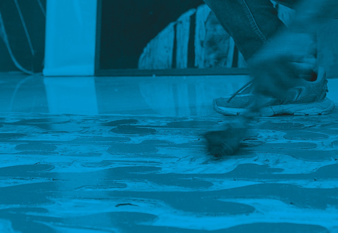 Descrição da imagem: fotografia horizontal colorida com filtro azul. Um detalhe de tênis diante de uma matriz de xilogravura deitada no chão. Uma mão em movimento espalha tinta sobre a matriz. Atrás dos pés, um pouco fora de foco, há um quadro retangular na vertical. Fim da descrição da imagem.
