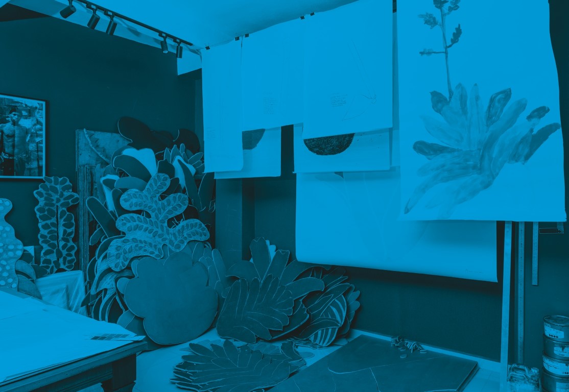Descrição da imagem: fotografia horizontal colorida com filtro azul. Ateliê de Santídio Pereira. No canto esquerdo da imagem há uma mesa e um retrato de Santídio pendurado na parede. No centro, há diversas matrizes grandes de xilogravura em formatos de flores e folhas, apoiadas na parede. À direita, há diversas pinturas em papel penduradas em um varal desde o teto, com desenhos de plantas e flores. Fim da descrição da imagem.