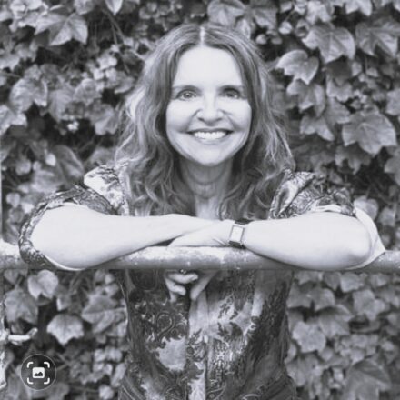 Imagem. Início da descrição de imagem: Retrato frontal em preto e branco de uma mulher sorridente com cabelos longos e ondulados. Ela está com os braços cruzados, apoiados sobre uma barra horizontal metálica em primeiro plano. Veste uma blusa com estampa de arabescos e usa um relógio de pulso no braço esquerdo. O fundo da imagem é composto por uma parede densa de folhagens de hera. Fim da descrição de imagem.