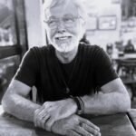 Imagem. Início da descrição de imagem: Retrato frontal em preto e branco de um homem de pele clara, com cabelos e barba brancos e curtos. Ele usa óculos de grau com armação clara e transparente, olha para o lado e sorri. Veste uma camiseta escura e usa diversos acessórios: um colar com pingente circular, anéis nos dedos das mãos e pulseiras de couro e metal no pulso esquerdo. O homem está com os braços cruzados sobre uma mesa de madeira em primeiro plano, e o fundo da imagem mostra o interior desfocado de um estabelecimento comercial. Fim da descrição de imagem.