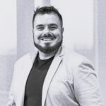 Imagem. Início da descrição de imagem: Retrato em preto e branco de um homem de barba cheia e bem cuidada, com cabelo escuro curto e espetado no topo. Ele sorri amplamente para a câmera, vestindo uma camiseta preta e um blazer cinza claro. O fundo é composto por faixas verticais claras e escuras desfocadas, possivelmente cortinas ou persianas. Fim da descrição de imagem.