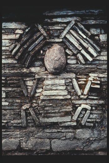 Descrição da imagem: Fotografia colorida de uma parede de pedra antiga com textura rústica. No centro, desenhado com as pedras, há um relevo formando um padrão geométrico com linhas e ângulos, na forma de uma pessoa sentada, ou de cócoras, vista de frente. Uma grande pedra oval está posicionada no meio do desenho, sugerindo um rosto. Na posição que estaria um chapéu, um padrão que lembra um par de assas pousa sobre a cabeça dessa figura. As pedras têm tons de cinza e marrom, com manchas e desgastes visíveis. Fim da descrição da imagem.