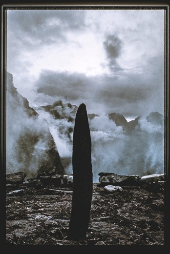 Descrição da imagem: Fotografia colorida com baixa saturação. No centro, uma estrutura vertical escura, que remete a um monolito, se ergue do chão de terra contra um fundo de névoa e vapor. Ao redor, rochas e terreno acidentado são visíveis. O céu está carregado de nuvens densas. A paisagem ao fundo são de montanhas recobertas de névoas. Fim da descrição da imagem.