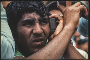 Descrição da imagem: Fotografia colorida horizontal. Retrato de um homem com expressão tensa. Ele segura um rádio de pilha próximo ao ouvido. Ele tem cabelos escuros e ligeiramente encaracolados, de pele parda. O fundo da imagem está desfocado. Fim da descrição da imagem.
