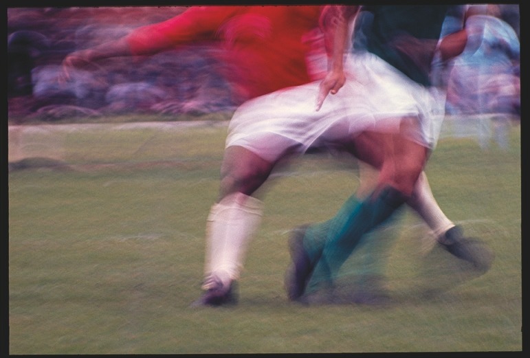 Descrição da imagem: Fotografia colorida horizontal. Imagem borrada de dois jogadores de futebol em movimento. Um jogador está vestindo uma camisa vermelha e calções brancos, enquanto o outro está usando uma camisa verde e calções brancos. Ambos estão em ação, com as pernas e corpos desfocados. O campo de futebol verde pode ser visto em segundo plano, junto com espectadores desfocados. Fim da descrição da imagem.