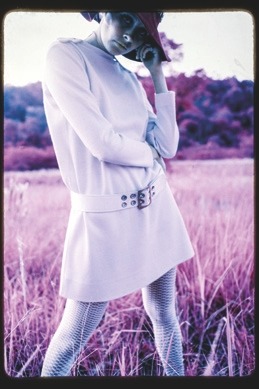 Descrição da imagem: Fotografia colorida vertical. A imagem tem efeito de cor alterado. Uma mulher está em pé em um campo. Ela veste um chapéu escuro, um casaco branco longo com fivela e meias-calças grossas. Sua postura é frontal, ligeiramente de perfil, com uma mão próxima ao rosto e outra na cintura. O campo ao redor tem uma coloração lilás, roxa e ocre. O céu e o fundo têm tons azulados. A imagem tem bordas escurecidas. Fim da descrição da imagem.
