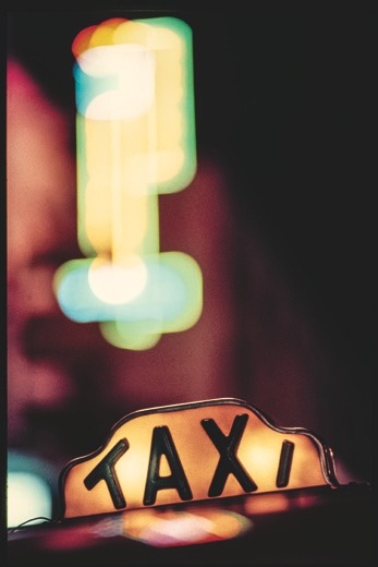 Descrição da imagem: Fotografia colorida vertical. Na parte inferior da imagem, um letreiro escrito "táxi" está iluminado com letras pretas sobre fundo amarelo. Acima dele, há uma luz desfocada e colorida, predominantemente verde e amarela, de outro letreiro em um prédio. O fundo é escuro, em cena noturna na cidade. Fim da descrição da imagem.