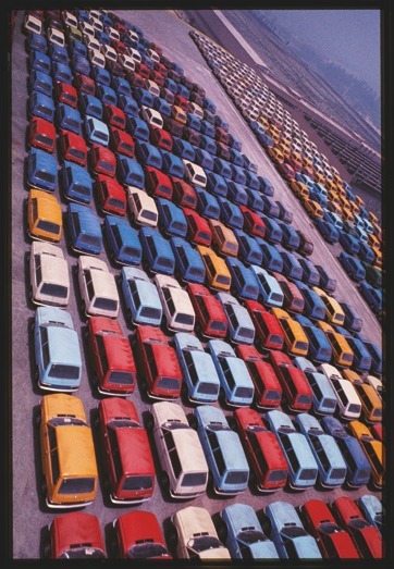 Descrição da imagem: Fotografia colorida vertical. Imagem de um grande estacionamento. Centenas de carros estão organizados em fileiras, criando diversos bolsões retangulares. Os veículos são de várias cores, principalmente azul, vermelho, branco e amarelo. A perspectiva da imagem cria uma sensação de profundidade, com as fileiras de carros se estendendo até o horizonte. O céu tem um tom acinzentado. A composição ressalta a escala do estacionamento. Fim da descrição da imagem.