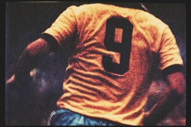 Descrição da imagem: Fotografia colorida horizontal. A imagem mostra um jogador de futebol da seleção brasileira, visto de costas, usando um uniforme amarelo com detalhes escuros nas mangas, com o número "9" destacado, e calção azul. A imagem captura o jogador em movimento em um recorte que mosta do pescoço até a cintura. Ao fundo, em desfoque, está a torcida no estádio. Fim da descrição da imagem.
