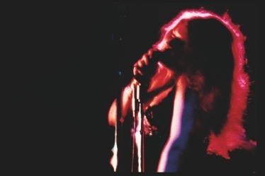 Descrição da imagem: Fotografia colorida horizontal. A imagem mostra uma cantora em uma performance ao vivo no palco. A imagem é escura, com iluminação dramática em tons de rosa e vermelho que realça o contorno do cabelo longo e esvoaçante. A artista está de perfil do lado dereito da imagem, voltado para esquerda. Segura o microfone em um pedestal perto da boca, com a cabeça inclinada e o ombro levemente flexionado para cima. Fim da descrição da imagem.