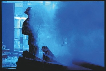 Descrição da imagem: Fotografia vertical colorida, em tons de azul, com uma cena de ambiente industrial. No centro da imagem, a silhueta de uma pessoa, provavelmente um trabalhador, usando capacete de proteção, é parcialmente coberta por uma densa nuvem de vapor . Ao fundo, estruturas metálicas e luzes difusas sugerem um cenário de fábrica ou indústria pesada. A fumaça ocupa grande parte da imagem, criando um efeito visual que encobre detalhes e acentua a atmosfera de trabalho intenso. Fim da descrição da imagem.