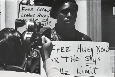 Descrição da imagem: Fotografia horizontal em preto e branco de uma manifestação. Em primeiro plano, um homem negro, com expressão séria, segura um cartaz com a inscrição "FREE HUEY NOW OR THE SKY'S THE LIMIT". À esquerda, uma pessoa de costas, com uma câmera fotográfica. Ao fundo, outro cartaz é visível, escrito "FREE BLACK LEADER HUEY". Fim da descrição da imagem.