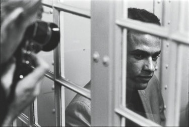 Descrição da Imagem: Fotografia horizontal em preto e branco de Huey Newton. Ele é um homem negro, está com expressão séria em está um ambiente fechado, atrás de uma divisória de vidro com estrutura metálica. À esquerda, há uma pessoa com uma câmera apontada para ele, do outro lado da divisória. Fim da descrição da imagem.
