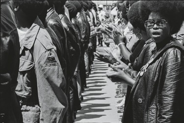 Descrição da Imagem: Fotografia horizontal em preto e branco do grupo de pessoas em pé, lado a lado, formando duas fileiras opostas. À direita, uma mulher negra, com cabelo black power e óculos, olha diretamente para a câmera, com uma expressão de atenção. As outras pessoas estão em de costas, batem palmas em protesto. Fim da descrição da imagem.