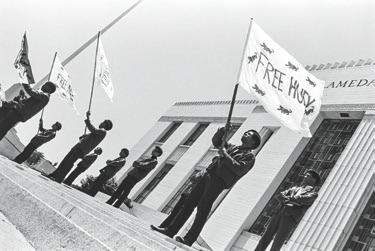 Descrição da Imagem: Fotografia horizontal em preto e branco mostrando uma manifestação em frente a um edifício com colunas. Várias pessoas, alinhadas em duas filas, seguram bandeiras com as palavras "Free Huey". Os manifestantes usam roupas escuras e boinas. Fim da descrição da imagem.