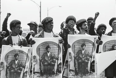 Descrição da Imagem: Fotografia horizontal em preto e branco de um grupo de manifestantes, homens negros, levantando o punho direito no ar. Eles seguram grandes cartazes com a imagem Huey Newton, ativista negro, que está sentado em uma cadeira de espaldar alto, vestindo jaqueta e segurando uma lança e um rifle, um em cada mão. Fim da descrição da imagem.
