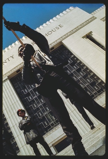 Descrição da imagem: Fotografia vertical colorida de dois homens em frente a um edifício de arquitetura clássica, com colunas altas e uma fachada de pedra clara, na qual se lê a palavra "COURT HOUSE". No primeiro plano, um homem em pé segura uma bandeira com ambas as mãos, usando uma boina preta, óculos de sol e um casaco de couro escuro. Em segundo plano, à esquerda, outro homem, também de pé, veste uma boina preta com detalhe em vermelho e um casaco longo preto, com as mãos cruzadas à frente do corpo. O céu azul claro preenche o fundo acima do edifício. Fim da descrição da imagem.