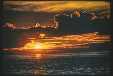 Descrição da imagem: Fotografia horizontal colorida de um pôr do sol sobre um rio. No centro da imagem, o sol parcialmente encoberto por nuvens densas ilumina o céu com tons de laranja, amarelo e dourado. Abaixo, o rio, que se assemelha a um oceano nesta imagem devido sua imensidão, reflete as cores do pôr do sol, com ondas suaves e sombras profundas. As nuvens formam silhuetas escuras no topo da imagem, criando um contraste de contra-luz. Fim da descrição da imagem.