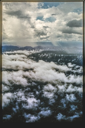 Descrição da imagem: Fotografia vertical colorida da uma paisagem aérea da floresta amazônica. A imagem mostra uma extensa cobertura de árvores vistas de cima, com muitas nuvens brancas pairando sobre a floresta. O céu ao fundo é coberto por nuvens cinzentas e brancas, com alguns raios de luz solar penetrando entre elas, iluminando partes da vegetação abaixo. As montanhas distantes aparecem suavemente ao fundo. Fim da descrição da imagem.
