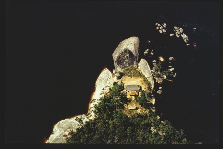 Descrição da imagem: Fotografia horizontal colorida do que aparenta ser uma pequena ilha rochosa, um uma ponta de península, vista de cima, cercada por água escura. A ilha possui uma vegetação densa na parte inferior, três construções pequenas e uma grande de habitação coletiva com teto de palha, perto de um grande rochedo vertical proeminente ao centro. À direita da imagem, há pedras menores espalhadas na água. O contraste entre a vegetação verde e o fundo de água escura realça a isolação da ilha. Fim da descrição da imagem.