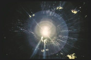 Descrição da imagem: Fotografia horizontal colorida de um feixe de luz intensa no centro da imagem, com raios de luz que emanam em várias direções. Ao redor, o fundo é predominantemente escuro, com algumas folhagens iluminadas pela luz, criando um efeito de brilho e difusão de luz, em uma cena capturada em direção a fonte de luz. Fim da descrição da imagem.