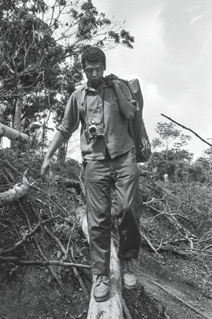 Descrição da imagem: fotografia em preto e branco na horizontal. Ele tem cabelo curto e escuro, usa camisa de manga comprida dobradas até os cotovelos, calça comprida, sapato e carrega uma câmera no pescoço e uma mochila nas costas. Está em pé em um tronco de árvore caído e olha para baixo. Ao fundo, a vegetação da floresta com inúmeras árvores secas e gravetos caídos. Fim da descrição da imagem.