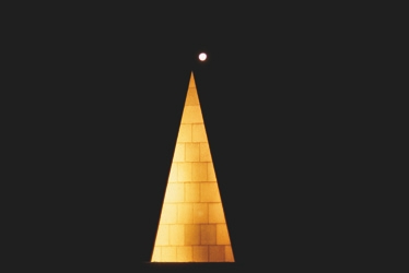 Descrição da imagem: Fotografia horizontal colorida de uma estrutura em forma de pirâmide iluminada em um fundo totalmente preto. A pirâmide, feita de blocos de pedra claros, é iluminada por uma luz dourada e, no topo, há uma pequena esfera luminosa, provavelmente uma lua cheia ou uma lâmpada, destacando-se no céu noturno. Fim da descrição da imagem.