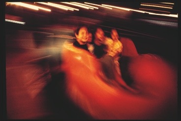 Descrição da imagem: Fotografia horizontal colorida de pessoas em movimento rápido, em um brinquedo giratório de parque de diversões. A imagem está desfocada e capturada com um efeito de longa exposição, criando um borrão de luzes e cores, especialmente em tons de vermelho e laranja. As pessoas ao centro aparecem sorrindo, mas seus rostos e corpos estão parcialmente distorcidos devido ao movimento. Fim da descrição da imagem.