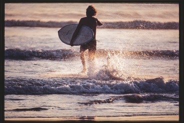 Descrição da imagem: Fotografia horizontal colorida de um surfista visto de costas, caminhando em direção ao mar com uma prancha de surfe embaixo do braço. A luz do pôr do sol cria um reflexo dourado na água enquanto ele avança pelas ondas rasas, com respingos ao seu redor. Fim da descrição da imagem.