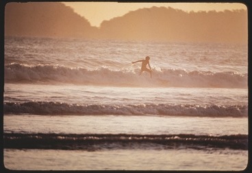 Descrição da imagem: Fotografia horizontal colorida de um surfista em ação, pegando uma onda no mar ao amanhecer ou entardecer. A luz dourada ilumina a cena, com o surfista se destacando em silhueta contra a água espumosa. Ao fundo, é possível ver o contorno de colinas ou montanhas distantes. Fim da descrição da imagem.
