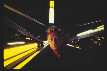 Descrição da imagem: Fotografia horizontal colorida de um homem de cabelos loiro claro olhando diretamente para a câmera, com um fundo escuro e várias luzes lineares coloridas. Sue rosot está centralizado e as luzes saem ao redor dele. As luzes são oriundas de lâmpadas fluoresecentes compridas, predominantemente em tons amarelos e brancos, e criam um efeito futurista a cena. Fim da descrição da imagem.