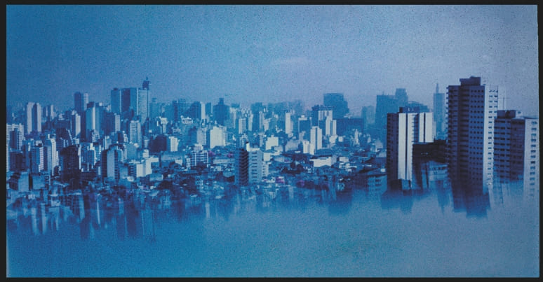 Descrição da imagem: Fotografia horizontal colorida de um skyline urbano em tons de azul, com vários edifícios de diferentes alturas e arquiteturas. A imagem ligeiramente refletida horizontalmente cria um efeito de duplicação e dá a impressão de que a cidade flutua em um fundo nebuloso ou aquoso. O tom geral da imagem é azulado devido a um efeito de de manipulação em sua revelação. Fim da descrição da imagem.