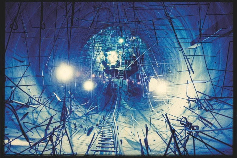 Descrição da imagem: Fotografia horizontal colorida de um túnel em construção. O túnel é iluminado por luzes artificiais, criando um tom azulado em toda a cena. Vários andaimes, fios e estruturas de metal estão espalhados pelo espaço, sugerindo um trabalho em andamento. Ao fundo, uma luz forte destaca operários e equipamentos em ação, adicionando profundidade à imagem. Fim da descrição da imagem.