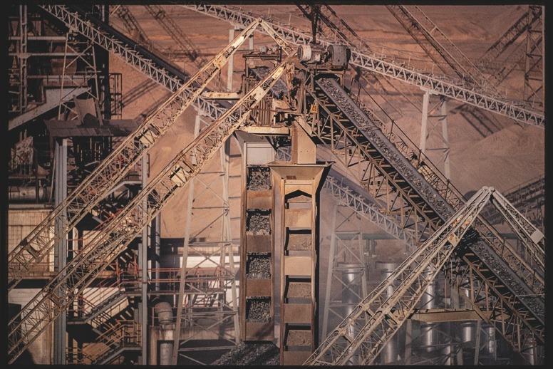 Descrição da imagem: Fotografia horizontal colorida de uma estrutura industrial complexa. A imagem mostra várias correias transportadoras inclinadas e plataformas de metal entrelaçadas em um ambiente de mineração, com montanhas de materiais a granel ao fundo. O tom geral é terroso, com detalhes metálicos e texturas de poeira, destacando a atividade industrial intensa. Fim da descrição da imagem.