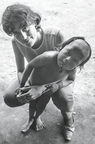 Descrição da imagem: fotografia em preto e branco de Claudia Andujar, agachada, com os braços envoltos na cintura de uma criança indígena. Claudia é uma mulher branca, de cabelos escuros, curtos e levemente ondulados. Usa regata clara, calça comprida e sapatilha. A criança está em pé, sem camisa e sem calça, tem o cabelo raspado até o topo da cabeça e comprido na parte de trás até a altura dos ombros e usa um adorno no queixo perto do lábio inferior. Fim da descrição da imagem.