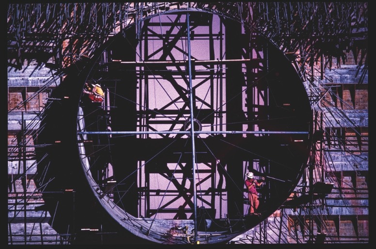 Descrição da imagem: Fotografia horizontal colorida de uma grande estrutura circular em construção. A imagem mostra a estrutura de um túnel ou reator, com andaimes e barras de aço ao redor. Dois trabalhadores, em roupas de segurança laranja e amarela, estão posicionados em diferentes pontos da estrutura, destacando a escala da construção e o trabalho humano envolvido. Fim da descrição da imagem.