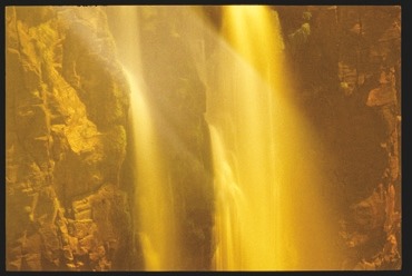 Descrição da imagem: Fotografia horizontal colorida de uma cachoeira iluminada por luzes em um tom douradas. A cachoeira é muito alta e a água cai suavemente entre as paredes rochosas, criando um efeito de véu translúcido, enquanto a iluminação realça os tons amarelados e dourados das pedras ao redor. Fim da descrição da imagem.