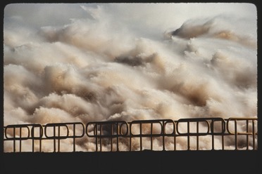 Descrição da imagem: Fotografia horizontal colorida de uma barreira de contenção diante de uma poderosa onda de água turbulenta. A água, em tons de marrom e branco devido à espuma e sedimentos, parece violenta e caótica, preenchendo quase todo o quadro da imagem. A barreira de metal em primeiro plano adiciona uma dimensão estrutural à cena natural intensa. Fim da descrição da imagem.