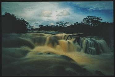 Descrição da imagem: Fotografia horizontal colorida de uma série de pequenas cachoeiras em um rio, cercadas por vegetação densa. A água, em tons de marrom claro, flui sobre as rochas, criando redemoinhos e espuma. O céu nublado ao fundo sugere um ambiente úmido e tropical. Fim da descrição da imagem.