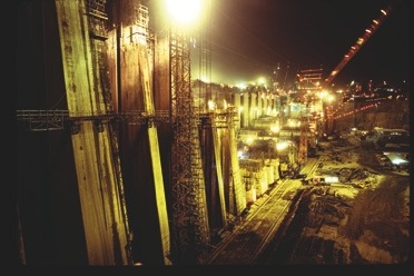 Descrição da imagem: Fotografia horizontal colorida de um grande canteiro de obras industrial durante a noite. A cena é iluminada por diversas luzes artificiais, destacando estruturas de concreto, gruas e andaimes. A imagem mostra uma ampla área de construção com intenso trabalho, evidenciado pela iluminação e equipamentos em operação. Fim da descrição da imagem.