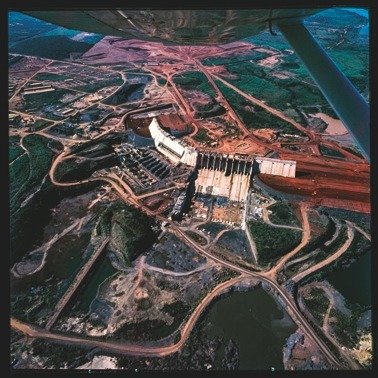 Descrição da imagem: Fotografia quadrada colorida de uma grande obra de construção vista de cima. A imagem mostra uma barragem em construção em meio a um terreno escavado, com caminhos de terra vermelha e áreas de vegetação ao redor. Várias estruturas de concreto estão em desenvolvimento, destacando a complexidade do projeto. A fotografia foi tirada de um avião, pois uma parte da asa é visível na parte superior da imagem. Fim da descrição da imagem.