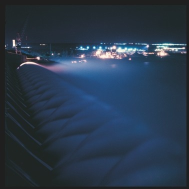 Descrição da imagem: Fotografia quadrada colorida de uma barragem durante a noite, iluminada por luzes artificiais. A imagem mostra uma série de estruturas semicirculares sobre a água, com uma névoa azulada cobrindo a superfície. Ao fundo, diversas luzes da instalação brilham na escuridão, em um ambiente de trabalho noturno de uma área industrial. Fim da descrição da imagem.