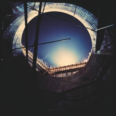 Descrição da imagem: Fotografia quadrada colorida de uma construção vista de dentro para fora, através de uma abertura circular em um túnel ou estrutura de concreto. A abertura revela o céu azul ao fundo, cercado por uma borda de metal e concreto. Algumas hastes metálicas atravessam o espaço, sugerindo que a obra está em andamento. A iluminação ao redor da abertura cria um efeito de contraste entre a escuridão interna e a luz exterior. Fim da descrição da imagem.