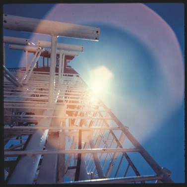 Descrição da imagem: Fotografia quadrada colorida de uma torre metálica capturada de baixo para cima, em direção ao sol. A estrutura de metal é composta por grades e plataformas, e a luz do sol cria um efeito de flare, com halos de luz coloridos ao redor da imagem. O céu é de um azul claro, e a perspectiva enfatiza a altura da torre. Fim da descrição da imagem.