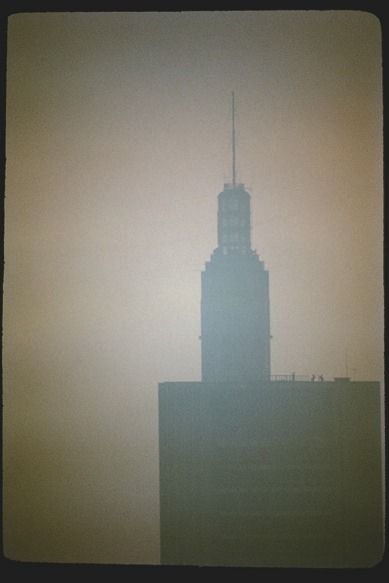 Descrição da imagem: Fotografia vertical colorida de um edifício alto, parcialmente encoberto por neblina ou poluição. A estrutura apresenta uma antena no topo e detalhes arquitetônicos no estilo de arranha-céus. A névoa densa suaviza os contornos da construção, que aparece em tons escuros contra um fundo alaranjado e esbranquiçado. Fim da descrição da imagem.